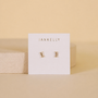A pair of Clear Baguette stud earrings by JaxKelly, hypoallergenic and gold plated, shown on a white brand card against a beige background.