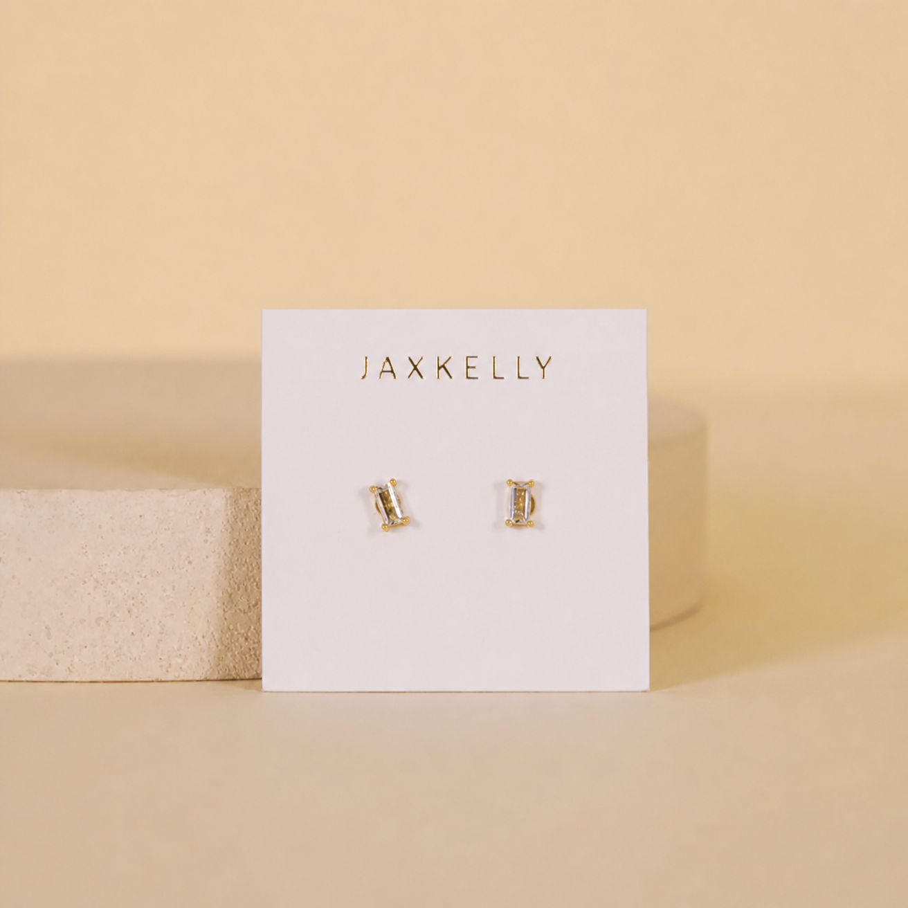 A pair of Clear Baguette stud earrings by JaxKelly, hypoallergenic and gold plated, shown on a white brand card against a beige background.