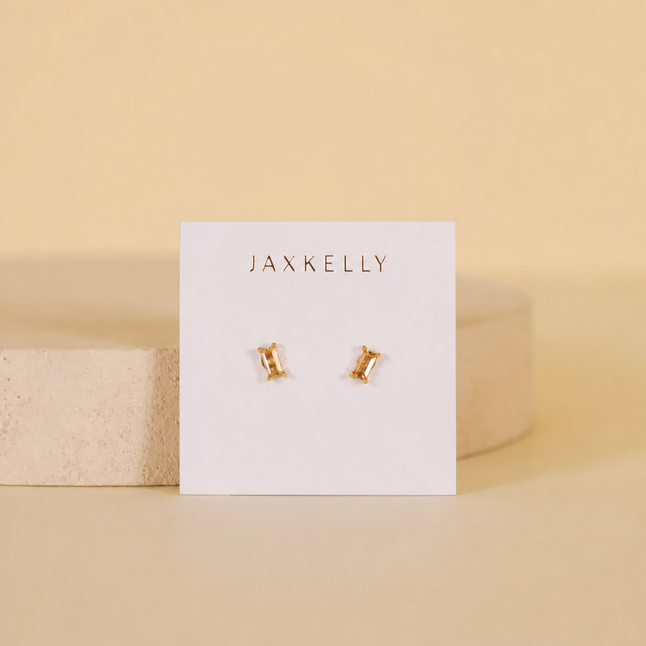 The Champagne Baguette stud earrings by JaxKelly feature rectangular baguette stones with 18k gold plating and sterling silver posts, shown on a white JaxKelly card against a beige background with a round stone prop.
