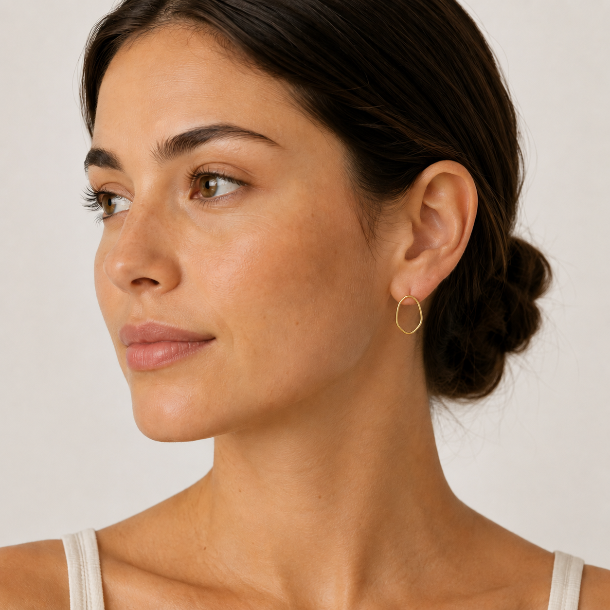 A woman with dark hair in a low bun wears JaxKelly’s Gold Organic Hoop earrings—handmade with 18k gold plating—and a light tank top, gazing to her left against a neutral background.