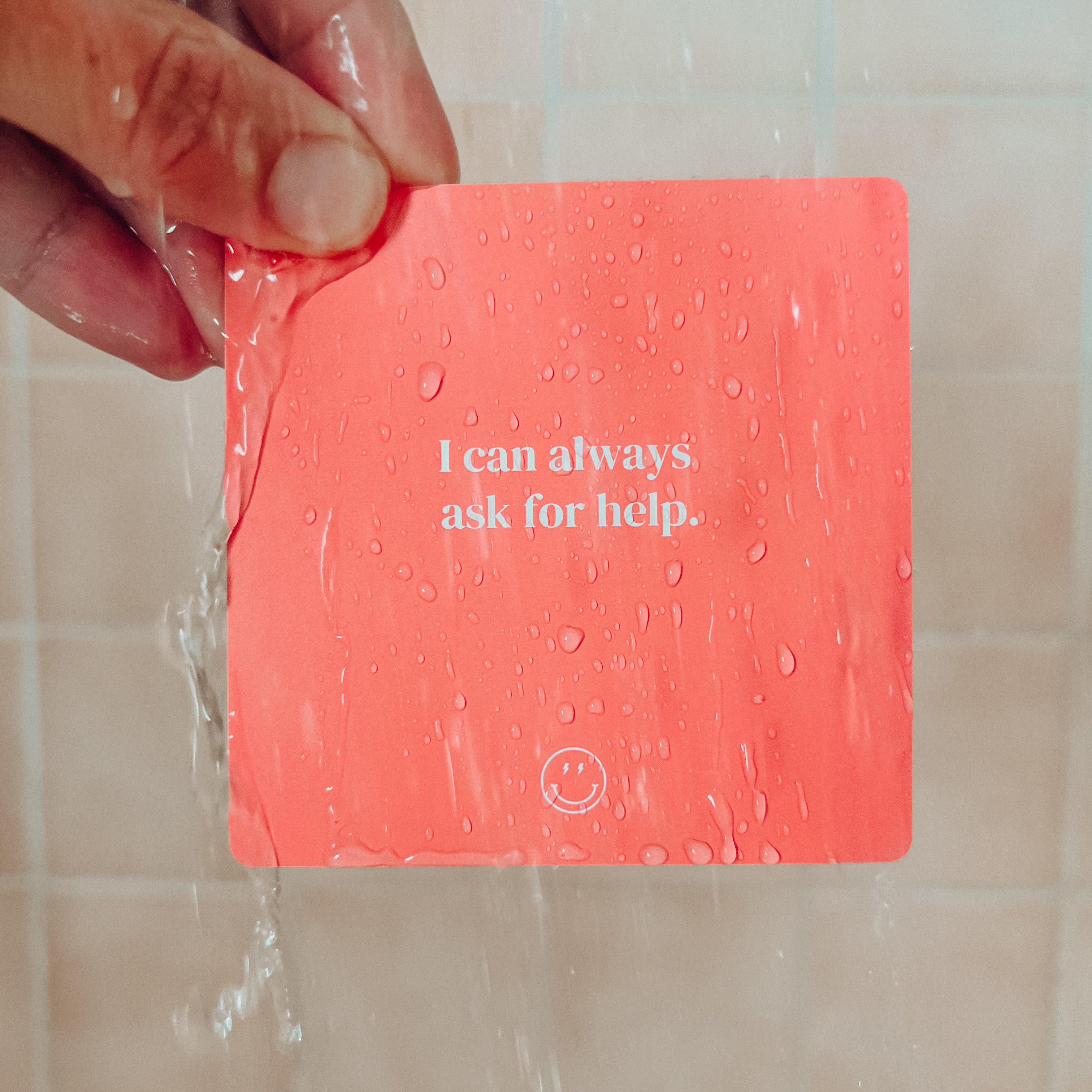 A hand holds a Teens Shower Affirmations™ card under running water. The waterproof red card, with “I can always ask for help” in white text and a smiley face, is ideal for sharing positive mindset quotes with teens.
