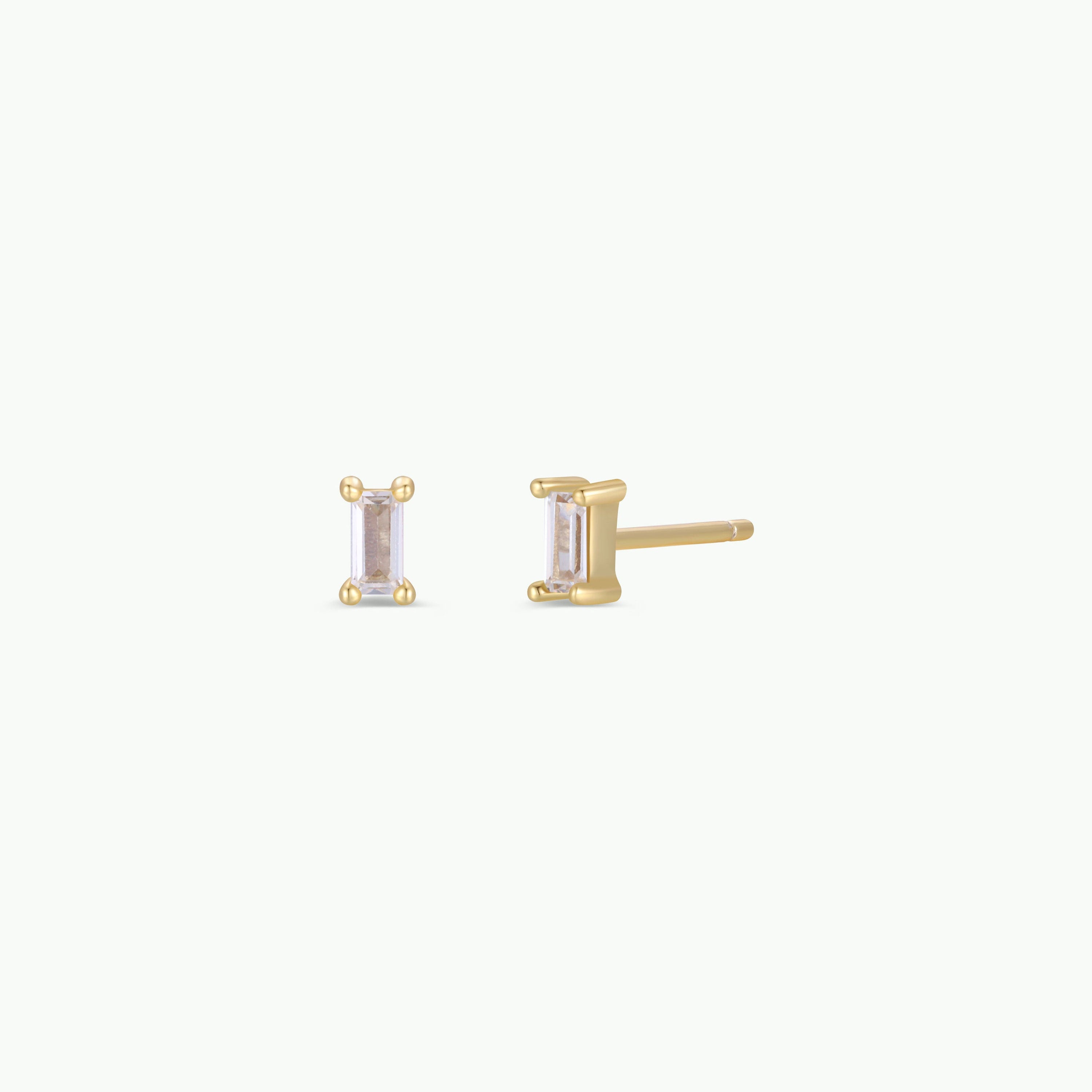 The Clear Baguette features two small baguette stud earrings with clear gemstones in yellow gold prongs and 18k gold plating, displayed front and side on a plain white background.