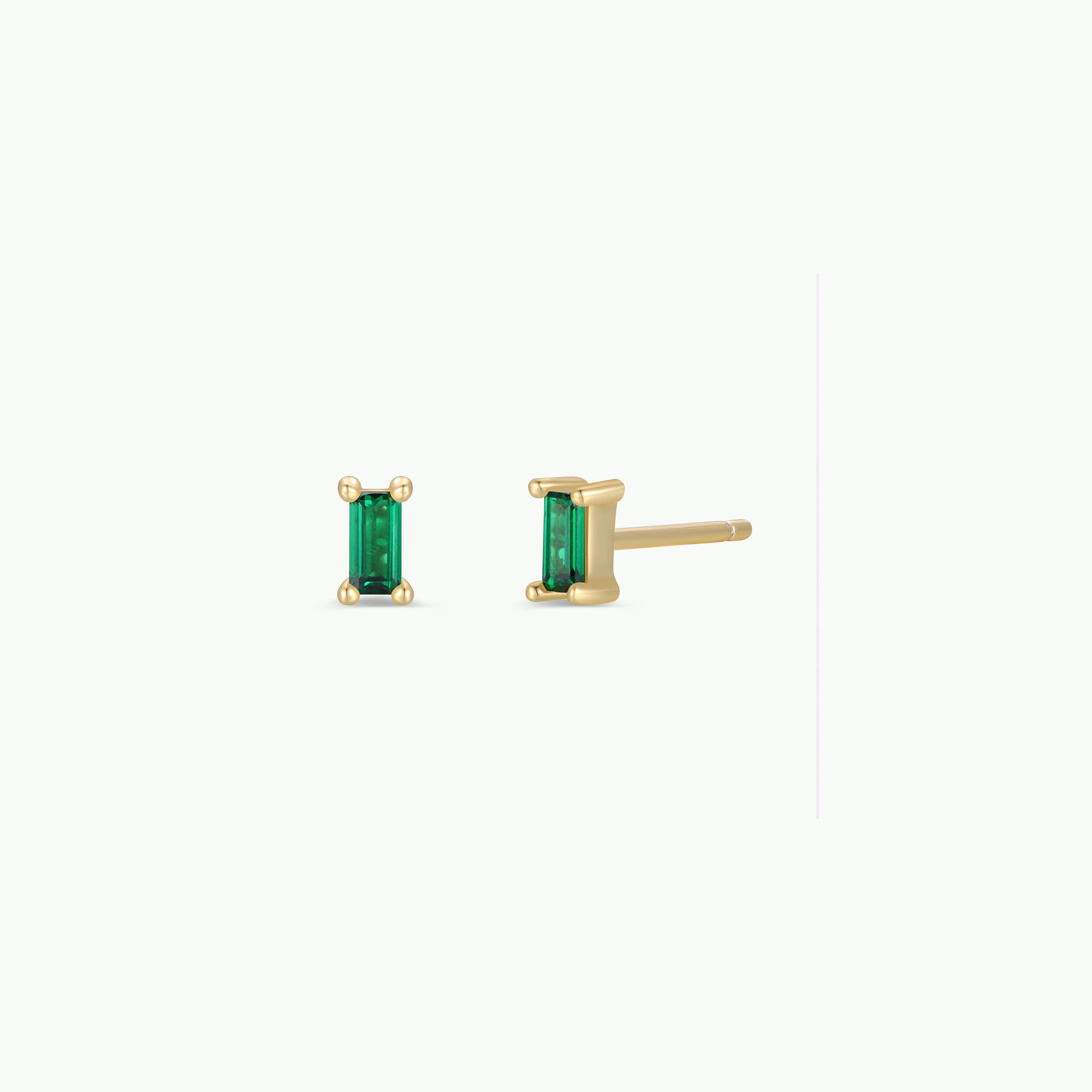 The Emerald Baguette features two gold-plated stud earrings, each showcasing a vertically set rectangular emerald-green gemstone with four prongs, displayed on a plain white background.