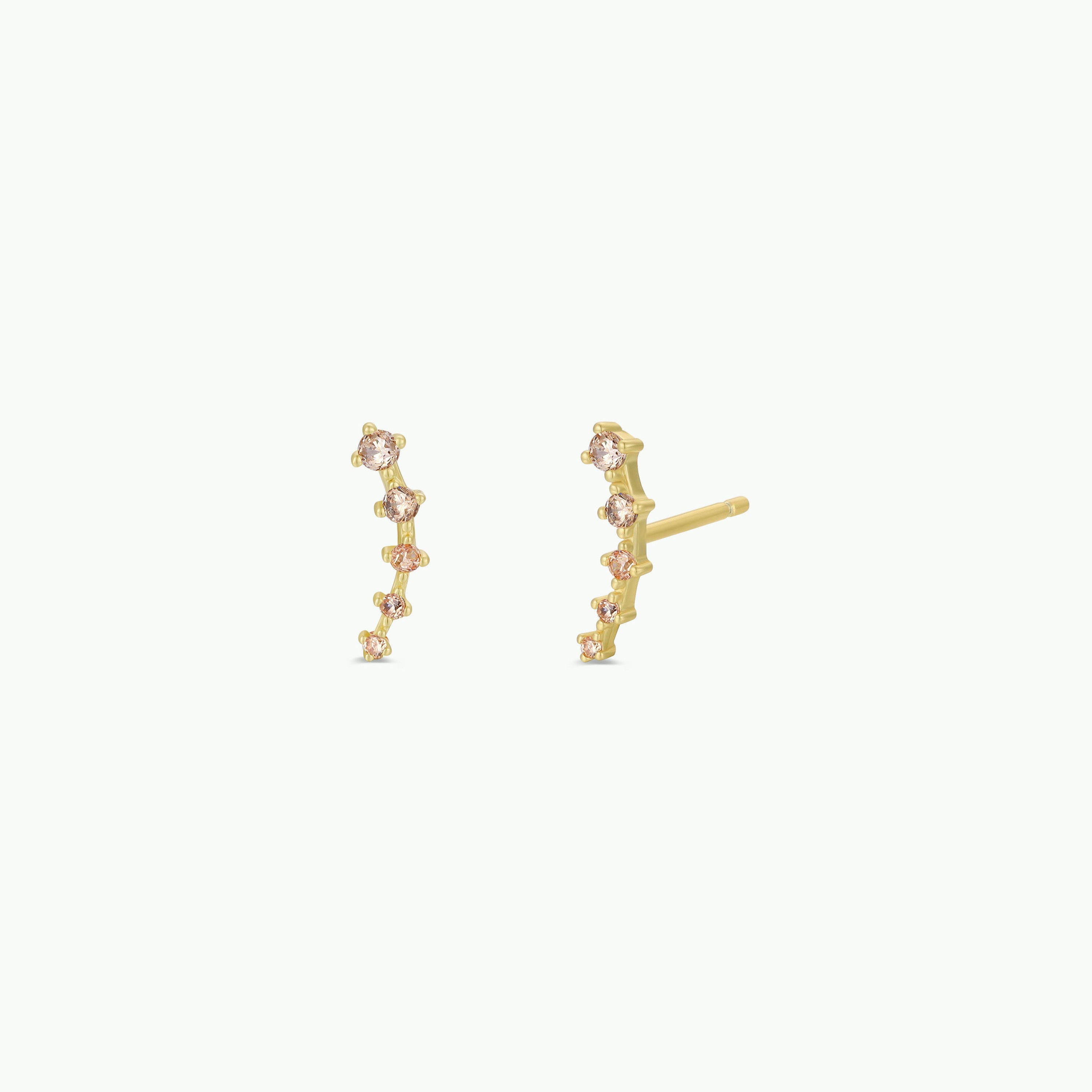 The Champagne Crawler features gold stud earrings with a curved line of six small, round sparkling gemstones set vertically, creating a delicate and elegant look. Displayed on a plain white background.