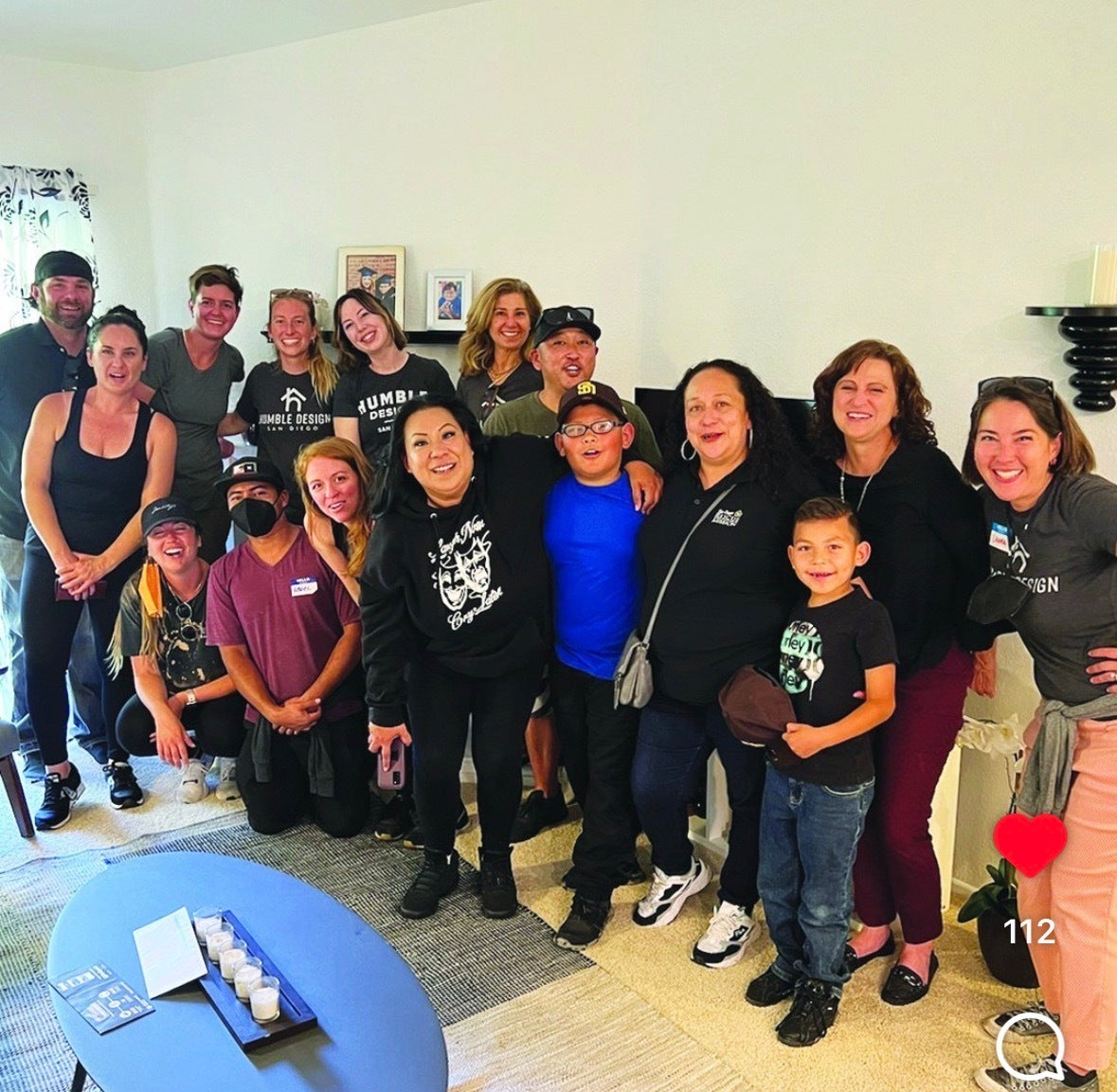 A group of sixteen people, including adults and children, smile and pose together in a living room. Some are kneeling, while others stand behind. A coffee table and house decor are visible in the foreground.