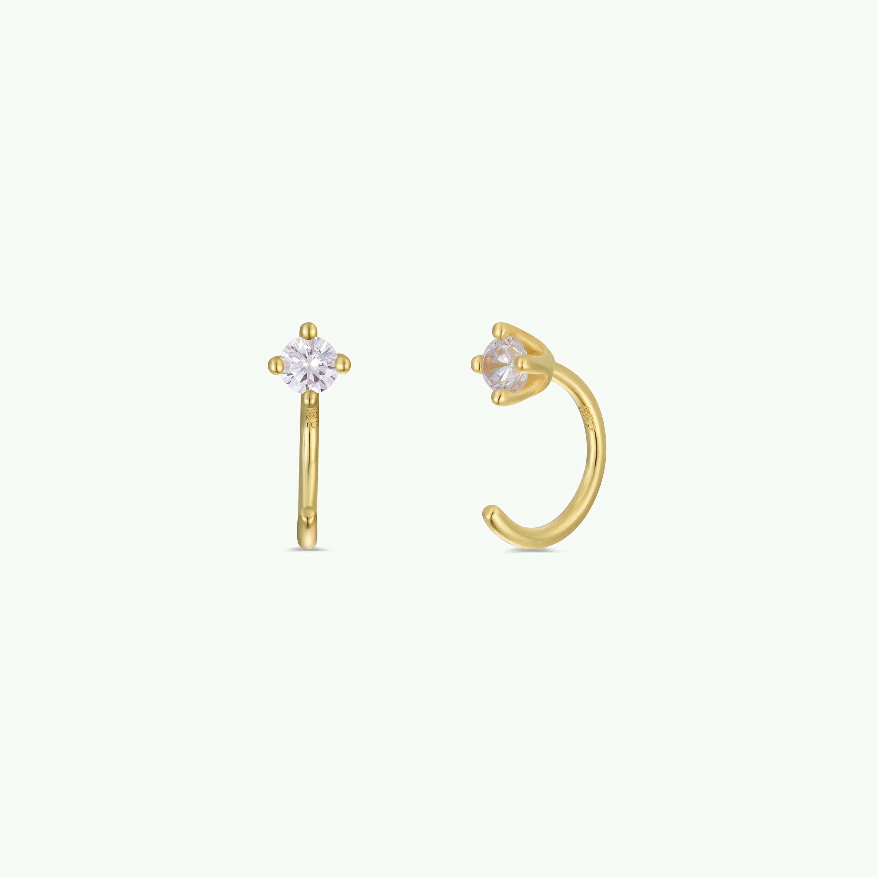 Two White CZ Huggie gold-plated earrings, each featuring a small round clear gemstone in a prong setting, are displayed on a plain white background—one shown from the front, the other from the side.