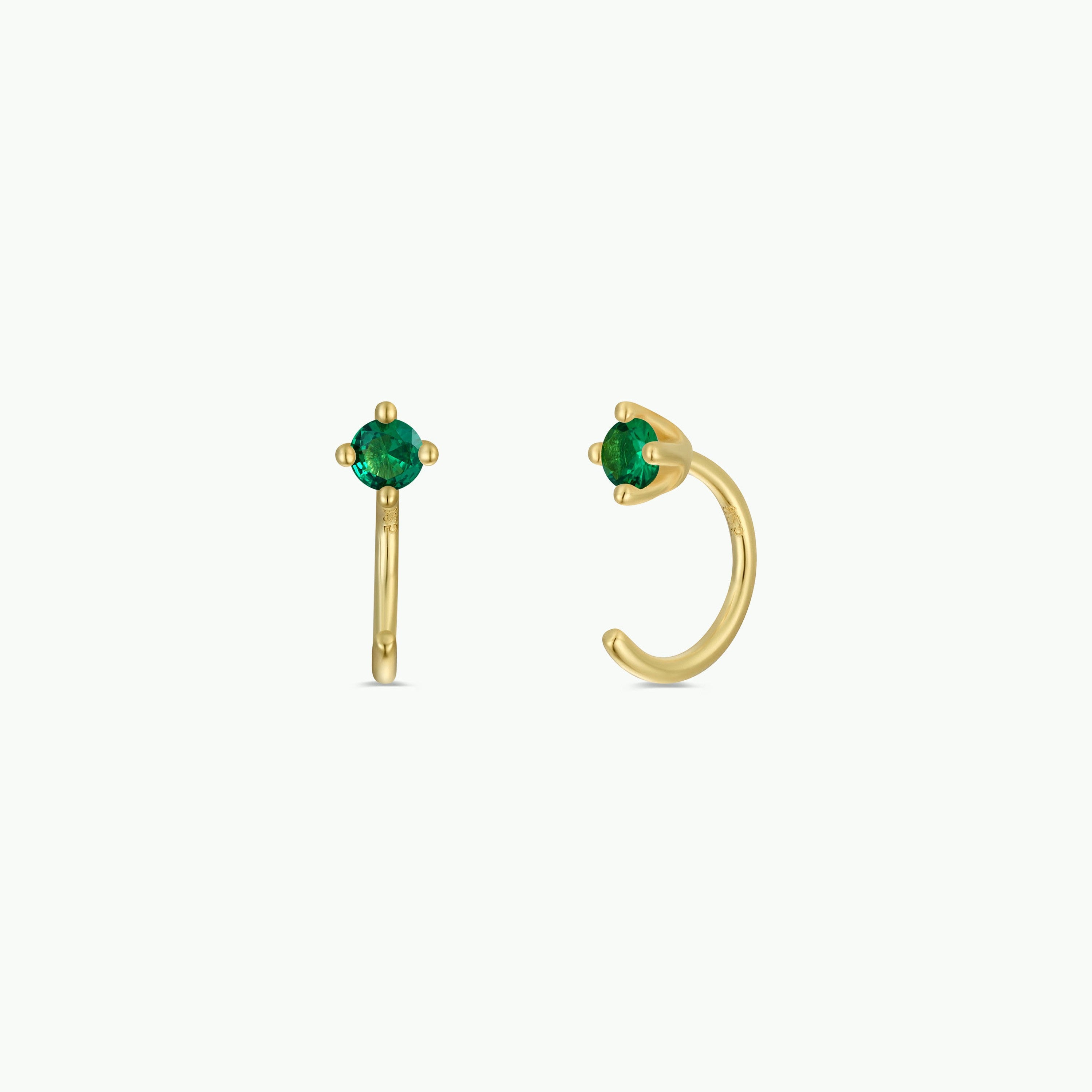 The Emerald CZ Huggie earrings feature round emerald CZ stones set at the center and are plated in 18k gold. They are showcased against a plain white background.