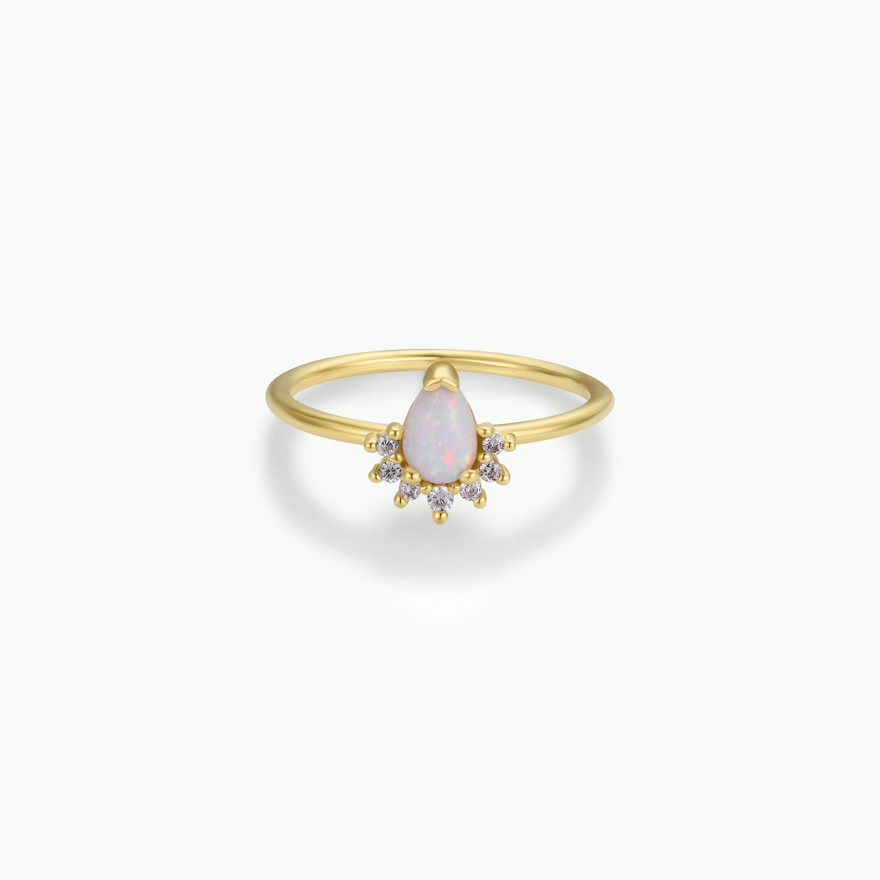 The Opal Burst Ring is an 18k gold-plated piece with a pear-shaped lab-grown Opal center, encircled by a halo of small round white diamonds, shown against a simple light background.
