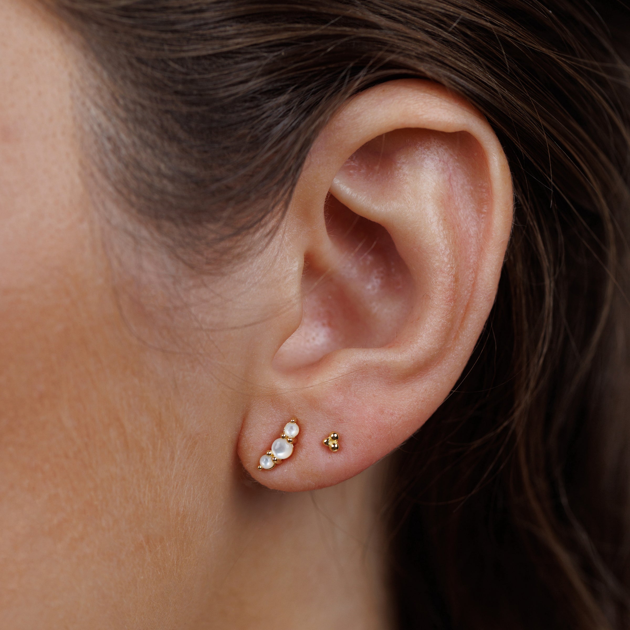 Close-up of a woman's ear wearing Crescent Gold Earrings - Pearl: a gold plated stud with three Mother of Pearls in a row, plus a delicate gold stud. Sterling silver posts offer comfort. Her brown hair is tucked back, and her skin appears smooth and natural.