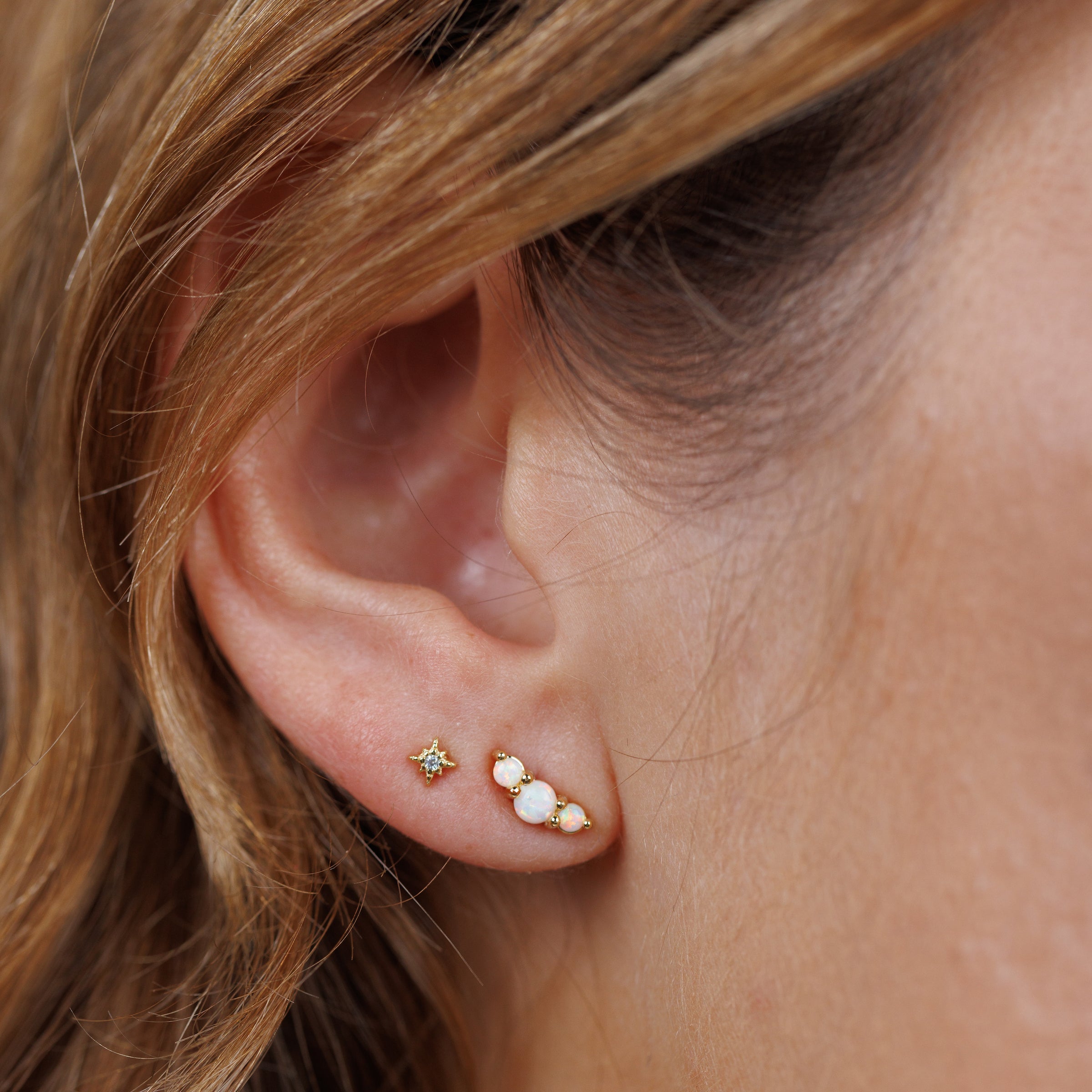 A woman’s ear adorned with Crescent Gold Earrings – White Opal, featuring three lab-grown white opals in a curved design alongside a delicate gold plated stud. Her brown hair softly frames her ear.