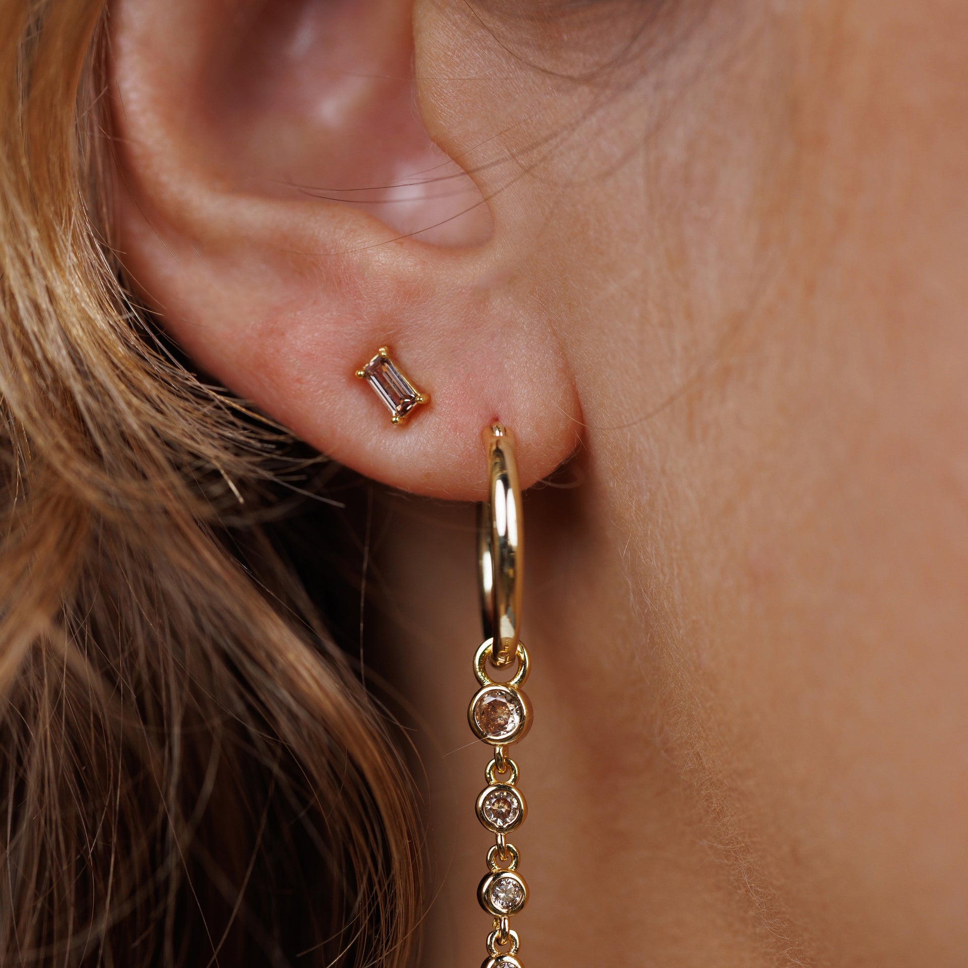 Close-up of a person’s ear wearing gold plated Coffee Baguette stud earring with sterling silver post in the upper lobe, and a gold hoop with four dangling round gemstones below. Strands of light brown hair are visible.