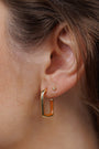 Close-up of a person’s ear with two earrings: a small round stud in the upper lobe and the Brown Enamel Large Rectangle Gold Hoop Earring in the lower lobe. Brown hair partially frames the ear.