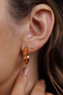 A close-up shows a woman’s ear with two 18k gold-plated earrings: a gold stud and the Vintage Hoop Earrings - Brown Enamel. Her nude-polished hand gently touches her earlobe, while her brown hair partially frames her ear.