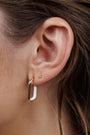 Close-up of an ear with two earrings: a small 18k gold plated stud in the upper lobe and a White Color Dipped Large Rectangle Hoop Earring in the lower lobe. Strands of brown hair are visible beside the ear.