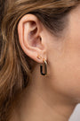 A close-up of a woman's ear shows two earrings: the Champagne Baguette gold-plated baguette stud with sterling silver post in the upper lobe and a larger gold and black rectangular hoop below. Brown hair is tucked behind her ear.
