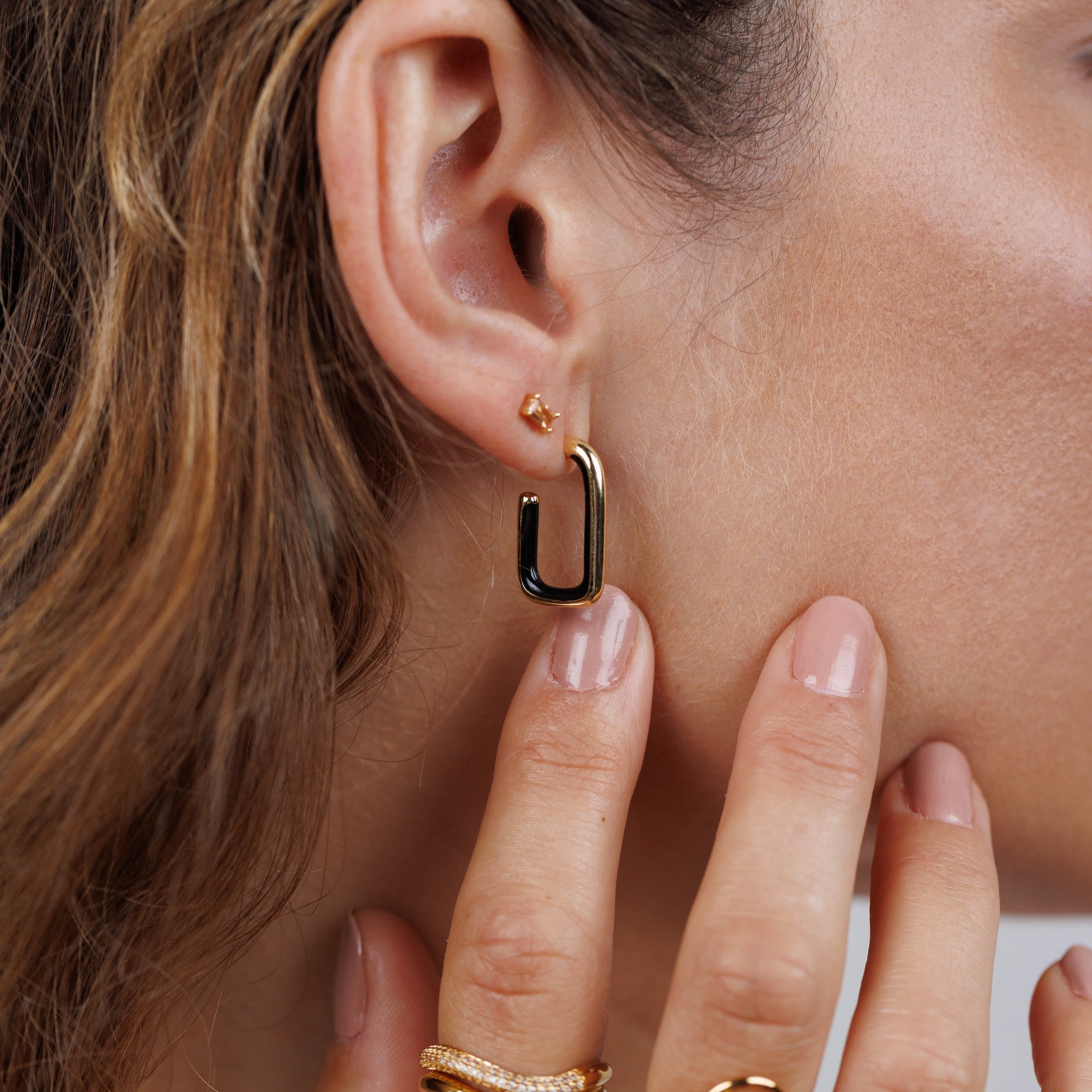 A close-up of a woman's ear with two earrings: a small gold stud and the Black Enamel Large Rectangle Hoop Earring. Her hand, with gold rings, touches her neck, and her nails are painted pale pink.