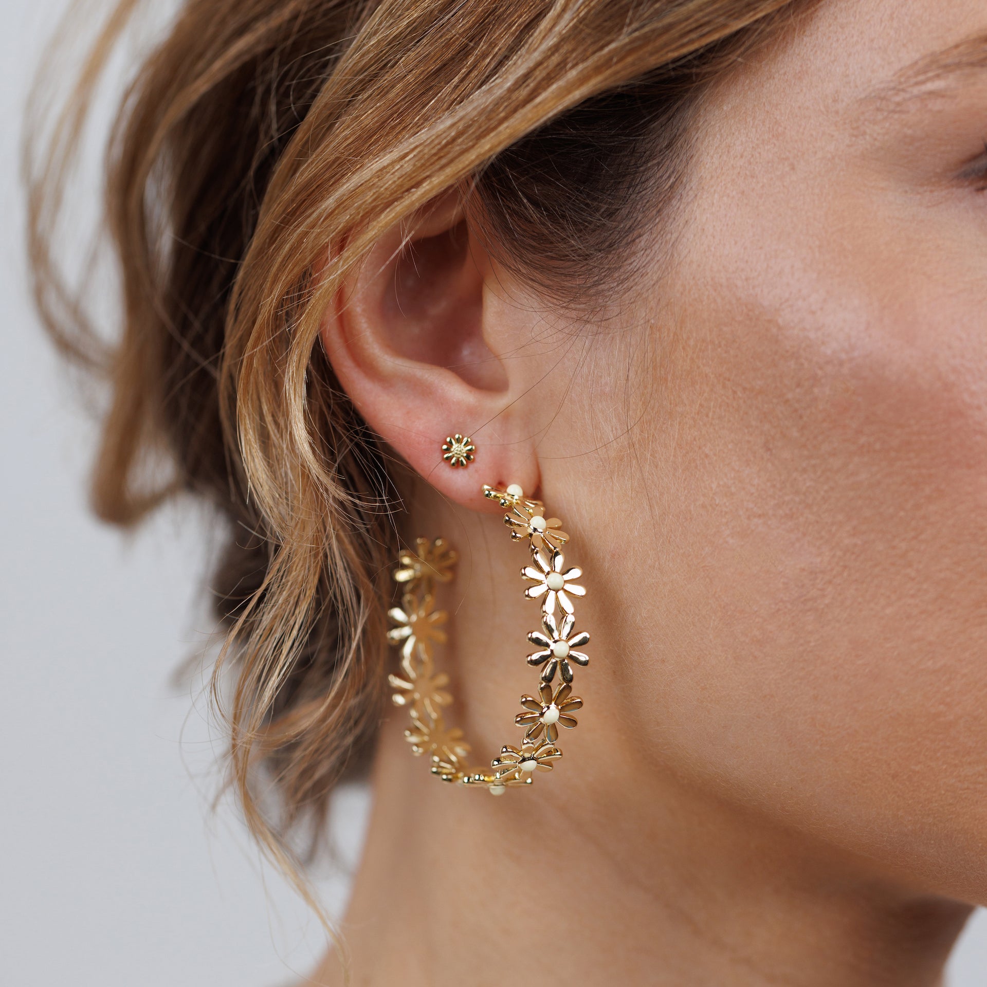 Close-up of a woman's ear wearing a large floral gold hoop earring, paired with the Petite Daisy Stud in an elegant ear stack. Part of her face and brown hair are visible.