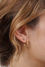 Close-up of a woman's ear with two gold earrings: a small stud on the upper lobe and the Petite Flower Hoop, a curved, textured gold earring with 18K gold plating and sterling silver posts, on the lower lobe. Light brown hair is visible nearby.
