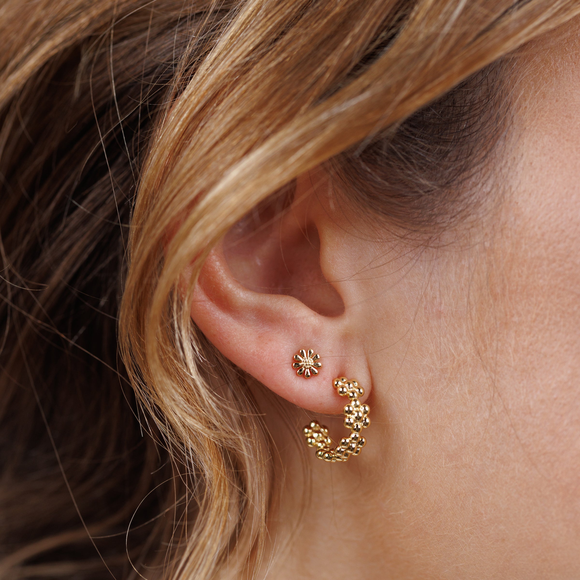 Close-up of a woman's ear with two gold earrings: a small stud on the upper lobe and the Petite Flower Hoop, a curved, textured gold earring with 18K gold plating and sterling silver posts, on the lower lobe. Light brown hair is visible nearby.
