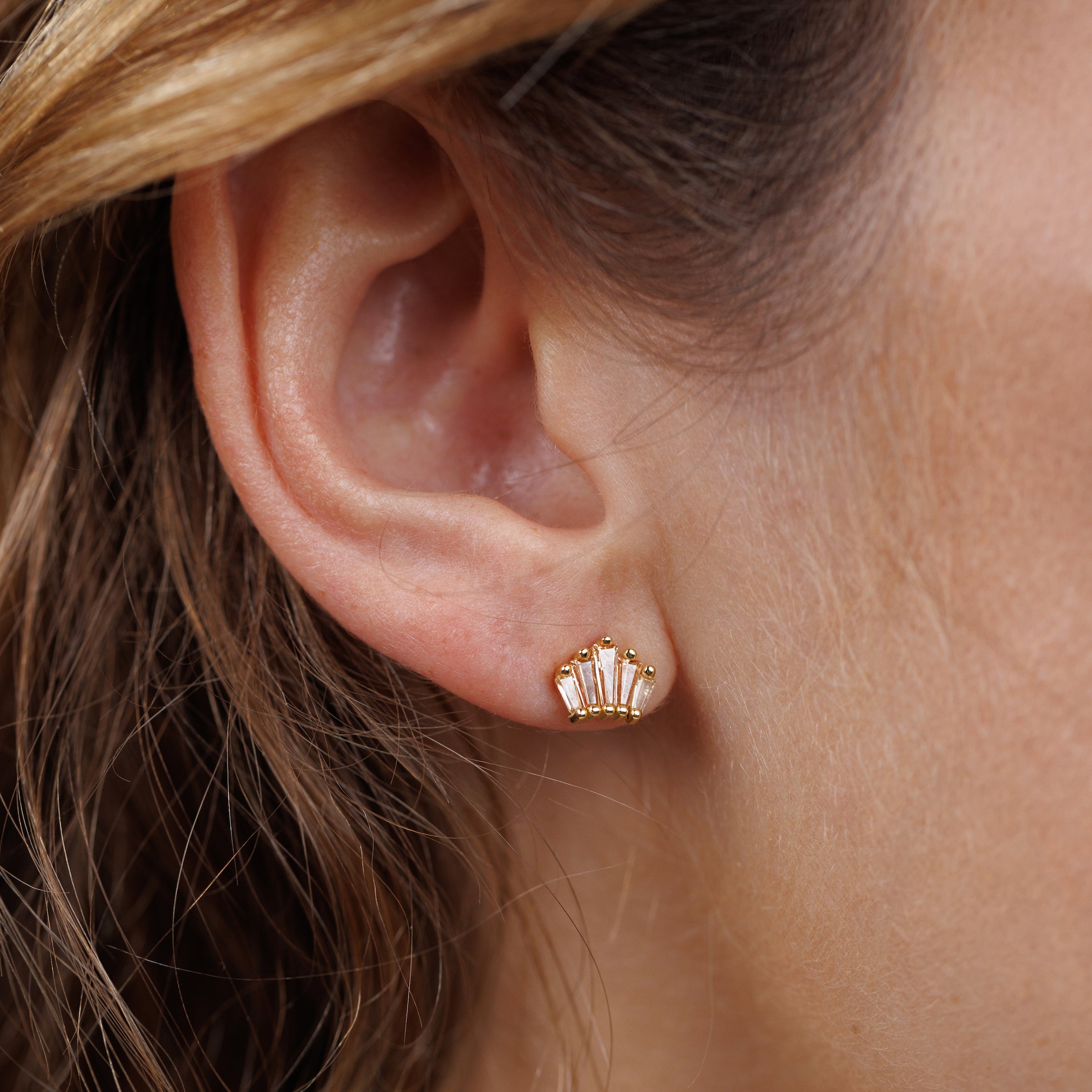 Close-up of a woman's ear with wavy brown hair, wearing JaxKelly's Champagne Baguette Crown Earrings—18K gold plated, tarnish-free studs featuring three clear rectangular gemstones in a fan shape.