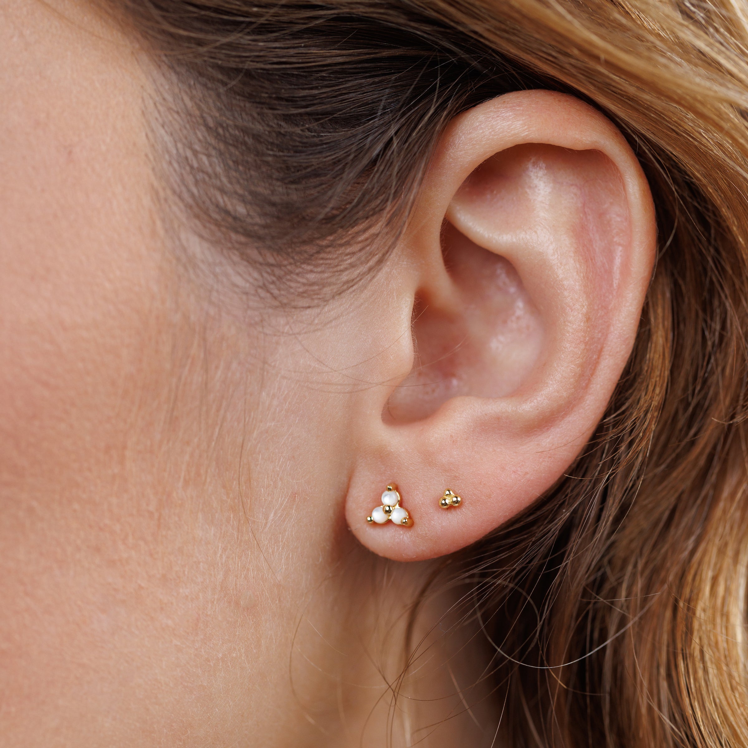 Close-up of a woman's ear adorned with Trio Cluster Gold Earrings - Pearl, featuring three clustered freshwater pearls on one stud and a tiny gold stud on the other, with her light brown hair tucked behind her ear.