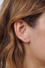 Close-up of a woman's ear adorned with the Trio Cluster Gold Earrings - Fire Opal, featuring gold-plated studs: one with a petite pink gemstone, the other showcasing three tiny shimmering fire opals framed by her wavy light brown hair.