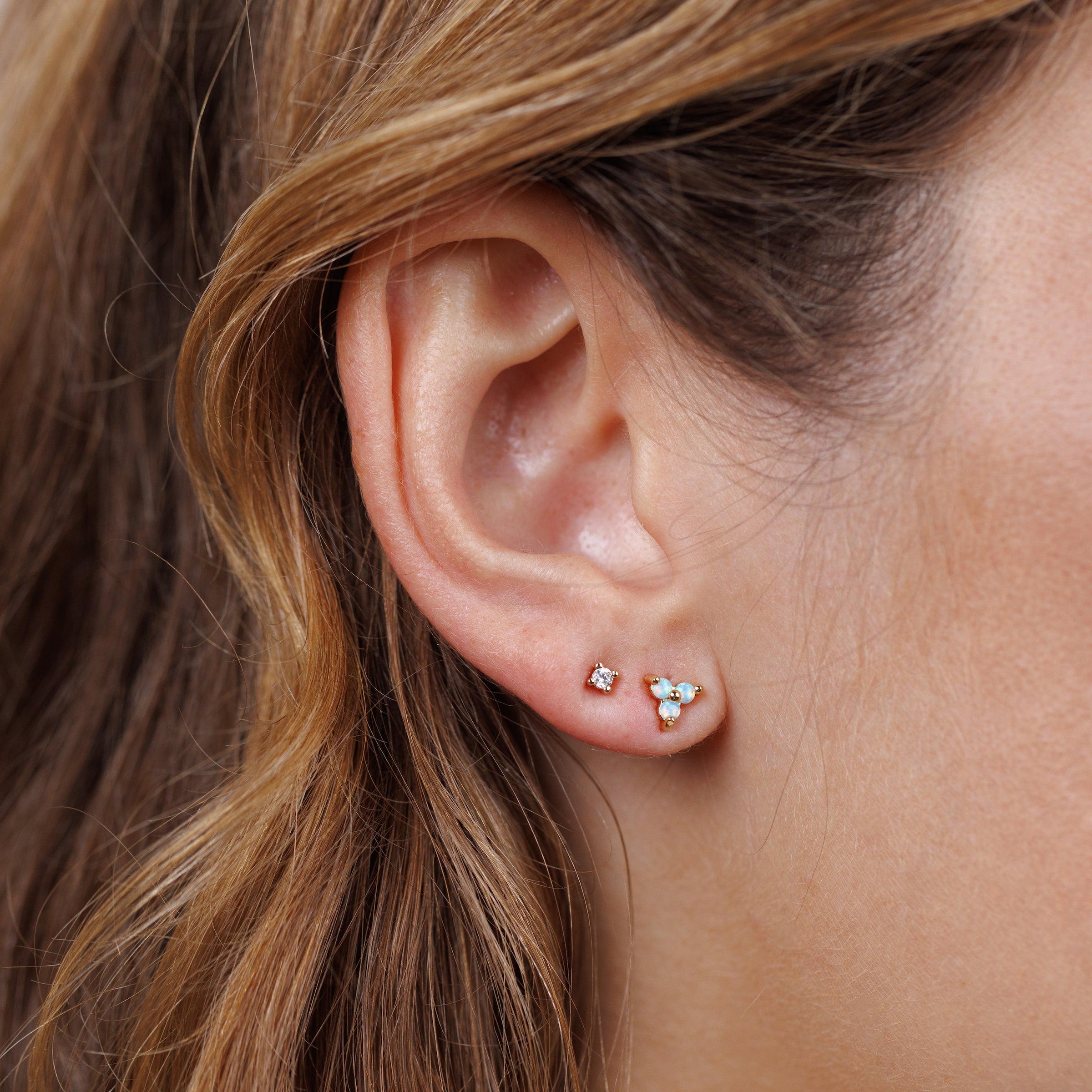 Close-up of a woman's ear adorned with the Trio Cluster Gold Earrings - Fire Opal, featuring gold-plated studs: one with a petite pink gemstone, the other showcasing three tiny shimmering fire opals framed by her wavy light brown hair.