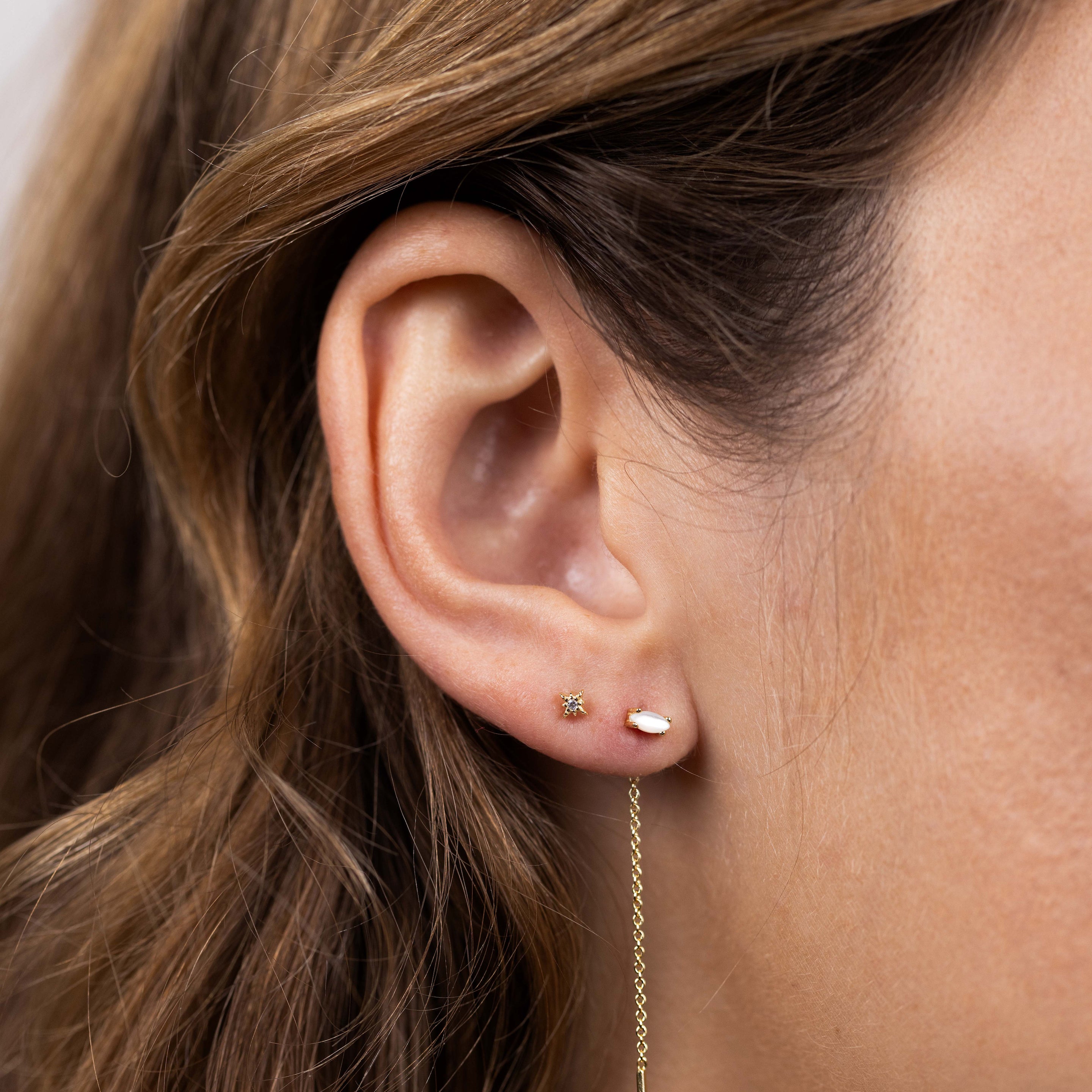 Close-up of a woman's ear wearing JaxKelly's Studded Threader - Mother of Pearl, along with a star-shaped stud and a small round stud. Her brown hair is tucked behind her ear as the threader earring dangles gracefully.