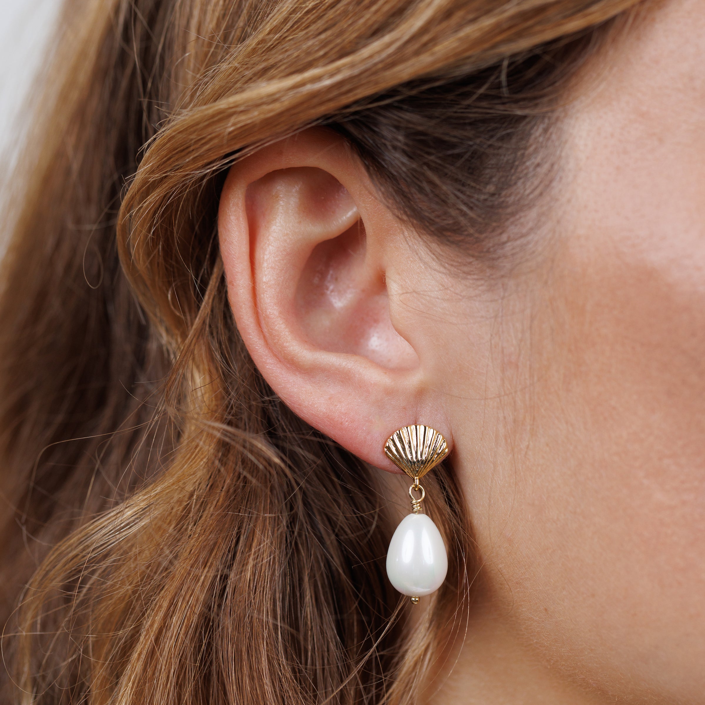 A close-up of a woman's ear wearing Seashell Pearl Drop Earrings, featuring 18K gold-plated seashells and a dangling freshwater pearl. Her light brown hair softly frames her face.