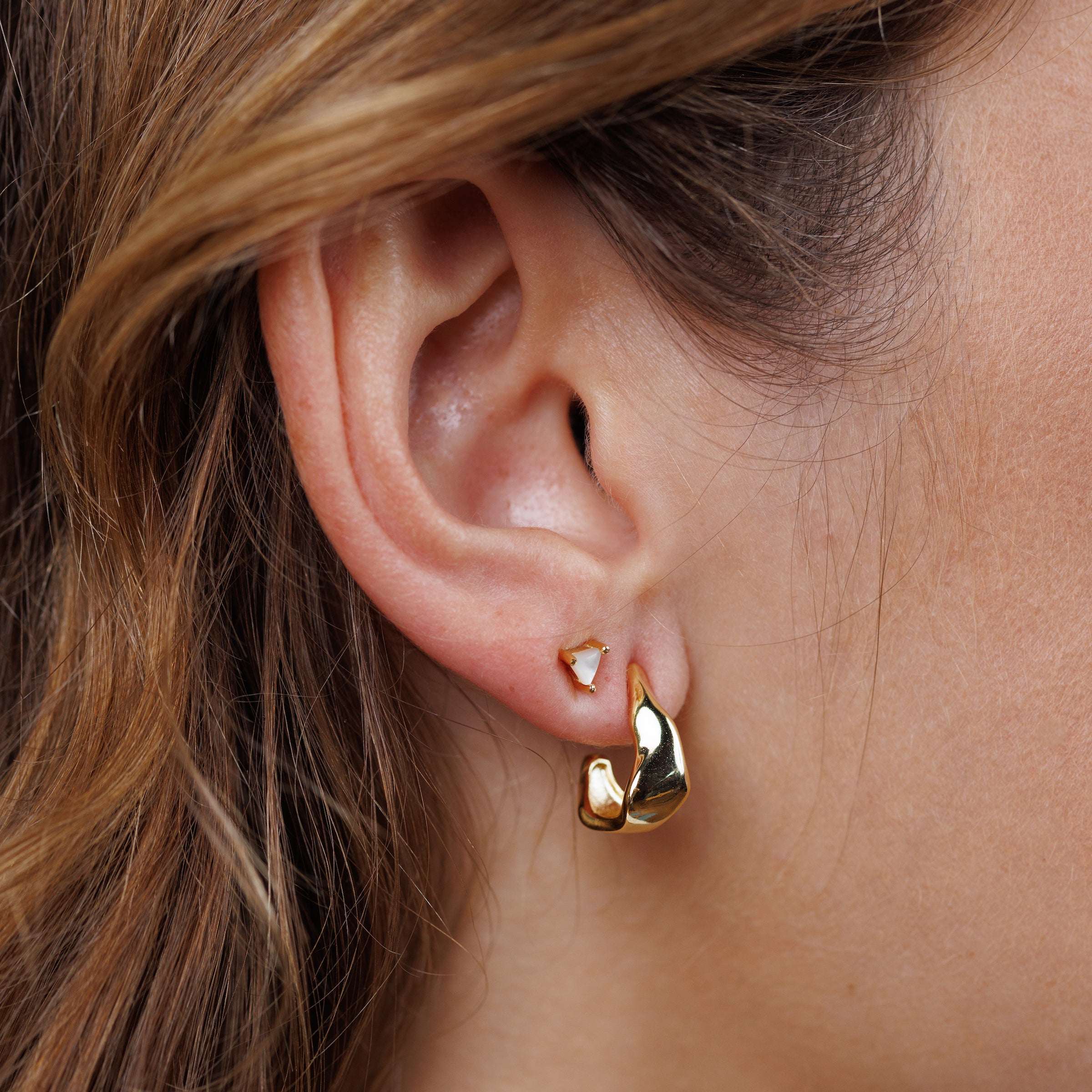 Close-up of a woman's ear with three gold earrings: a triangle stud, an abstract stud, and the Small Ripple Gold Hoop—featuring 18K gold plating and sterling silver posts. Her light brown hair is tucked behind her ear.