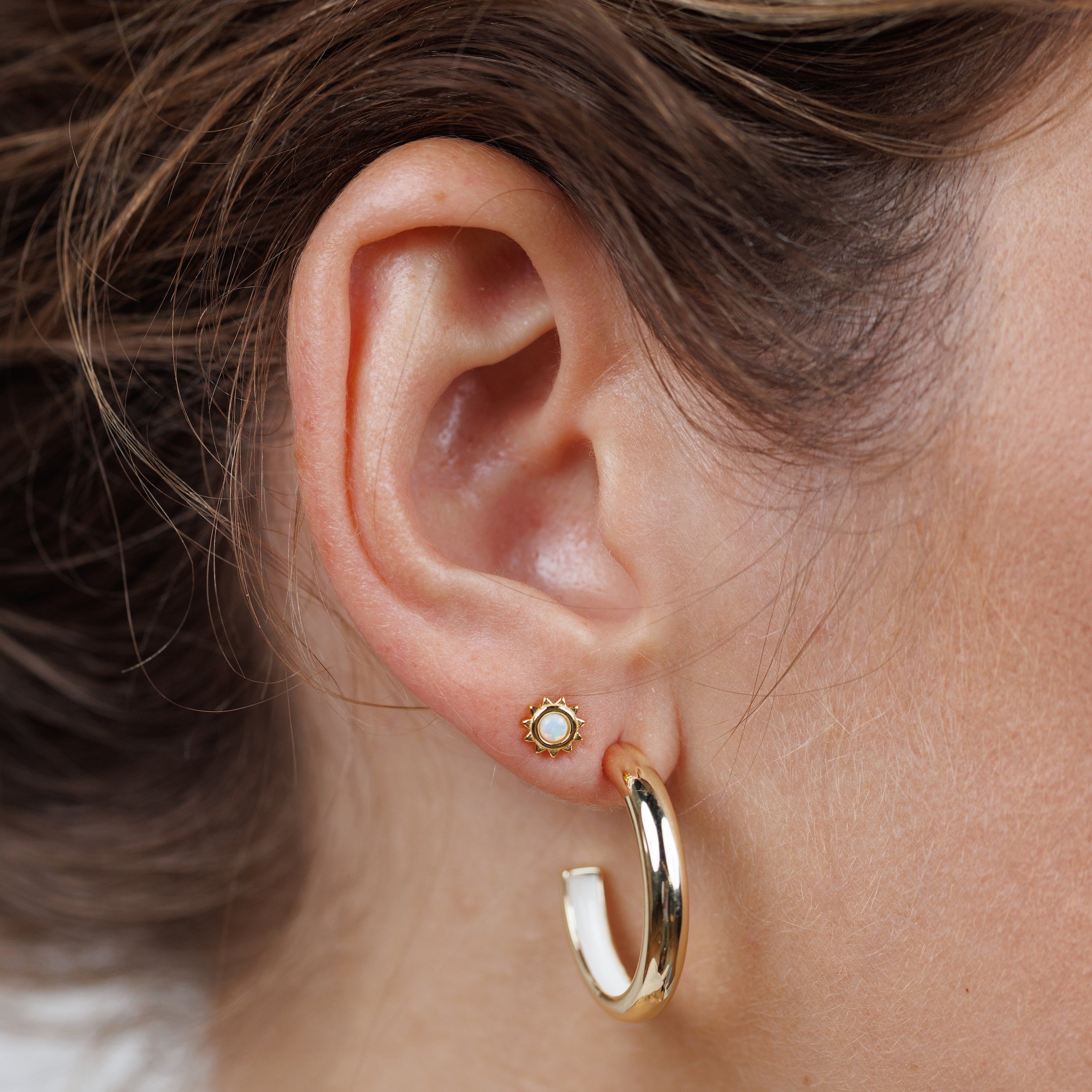 Close-up of a woman's ear wearing the White Opal Sun Stud, featuring a lab-grown white opal center with 18k gold plating in the upper lobe, and a larger gold hoop in the lower lobe. Hair is pulled back to showcase the earrings.
