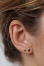 Close-up of a person’s ear with the Birthstone Stud Earring Set - May: one small round and one larger triangular emerald stud, both set in gold. Brown hair pulled back highlights these unique birthstone earrings.