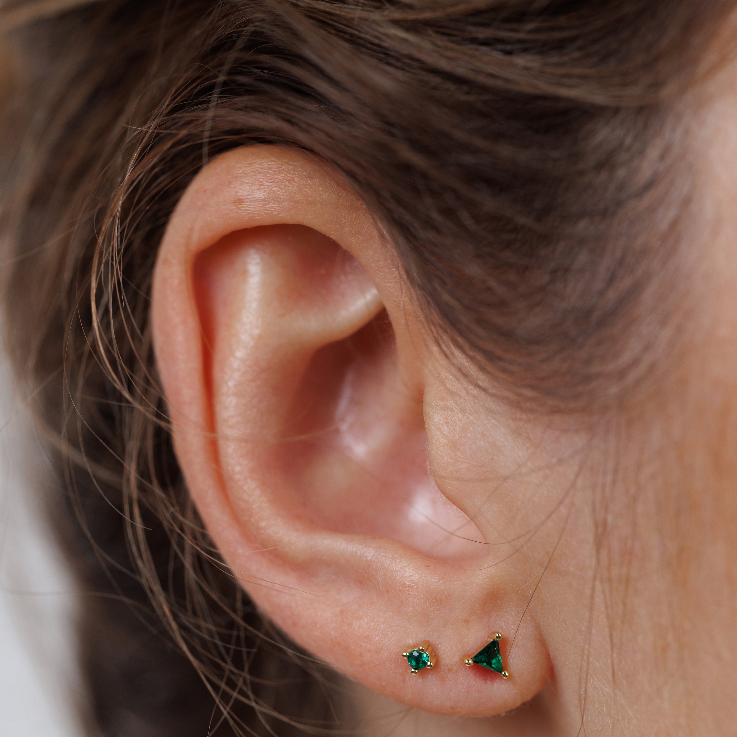 Close-up of a person’s ear with the Birthstone Stud Earring Set - May: one small round and one larger triangular emerald stud, both set in gold. Brown hair pulled back highlights these unique birthstone earrings.