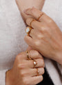 Close-up of two hands wearing several gold rings, including the Ring - Pave Wave - Champagne CZ, all shining with 18k gold plating against a soft, light-colored sweater.