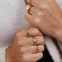 Hands adorned with multiple Wave Rings in 18k gold rest against the chest of someone wearing a soft, light sweater. Some rings showcase white stones and diamond-shaped designs.
