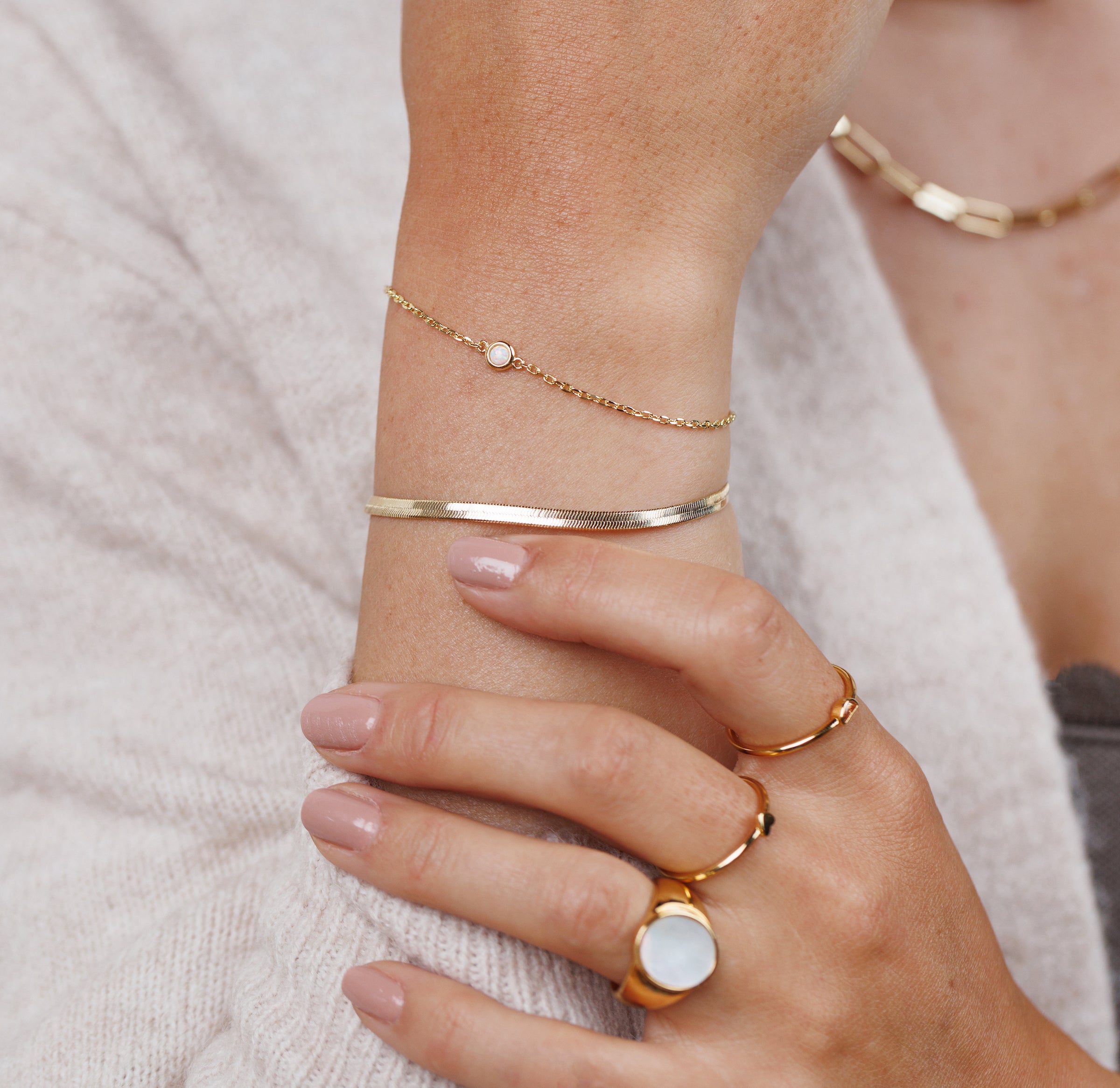 A close-up of a person’s hand wearing the Flat Snake Chain Bracelet, featuring delicate 18k gold plating and a small white stone, along with gold rings and a bangle. The person wears a light sweater and has neatly manicured nails.