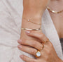 A close-up of a person’s hand wearing the Flat Snake Chain Bracelet, featuring delicate 18k gold plating and a small white stone, along with gold rings and a bangle. The person wears a light sweater and has neatly manicured nails.