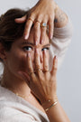 A woman with light brown hair poses with her hands framing her face, showcasing the Half Moon Champagne CZ Ring, along with stacking rings and neutral nail polish. She wears a beige top and looks directly at the camera.