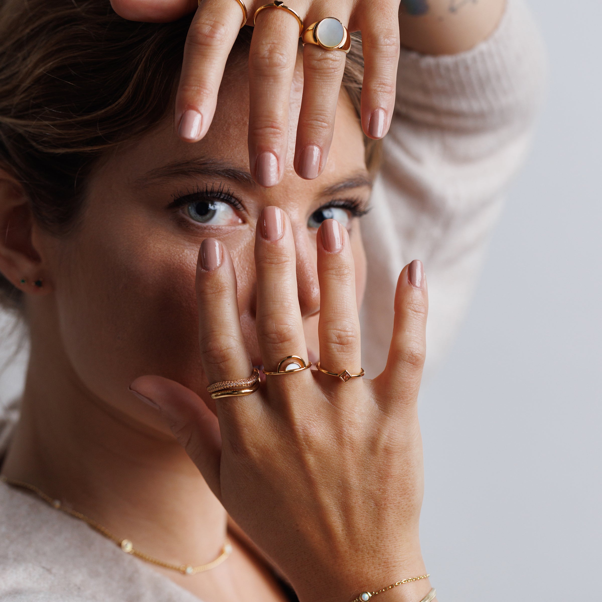 A woman with light brown hair poses with her hands framing her face, showcasing the Half Moon Champagne CZ Ring, along with stacking rings and neutral nail polish. She wears a beige top and looks directly at the camera.