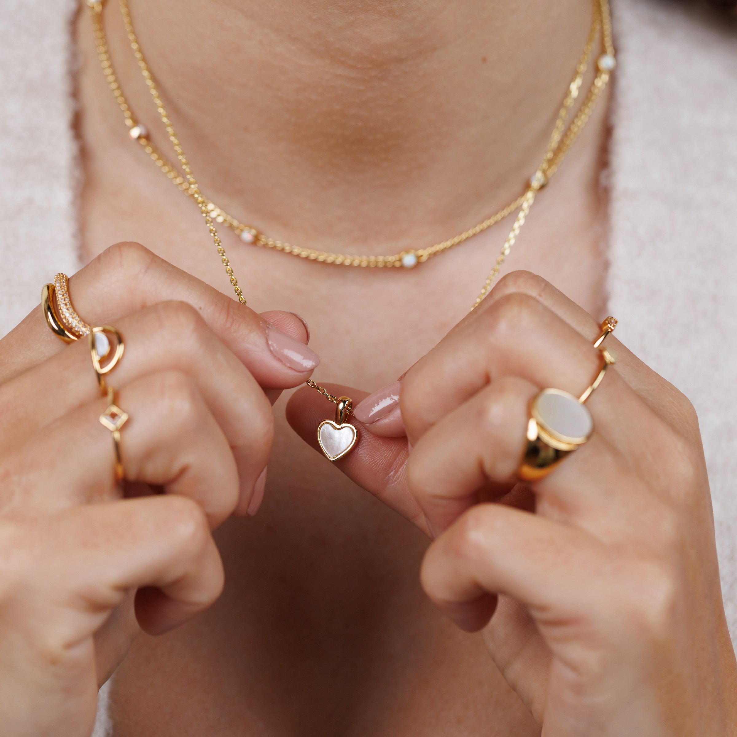 A person holds the Mother of Pearl Heart Pendant Necklace, wearing several gold rings and a layered necklace with small gems. The jewelry is the focus, while their face remains out of view.