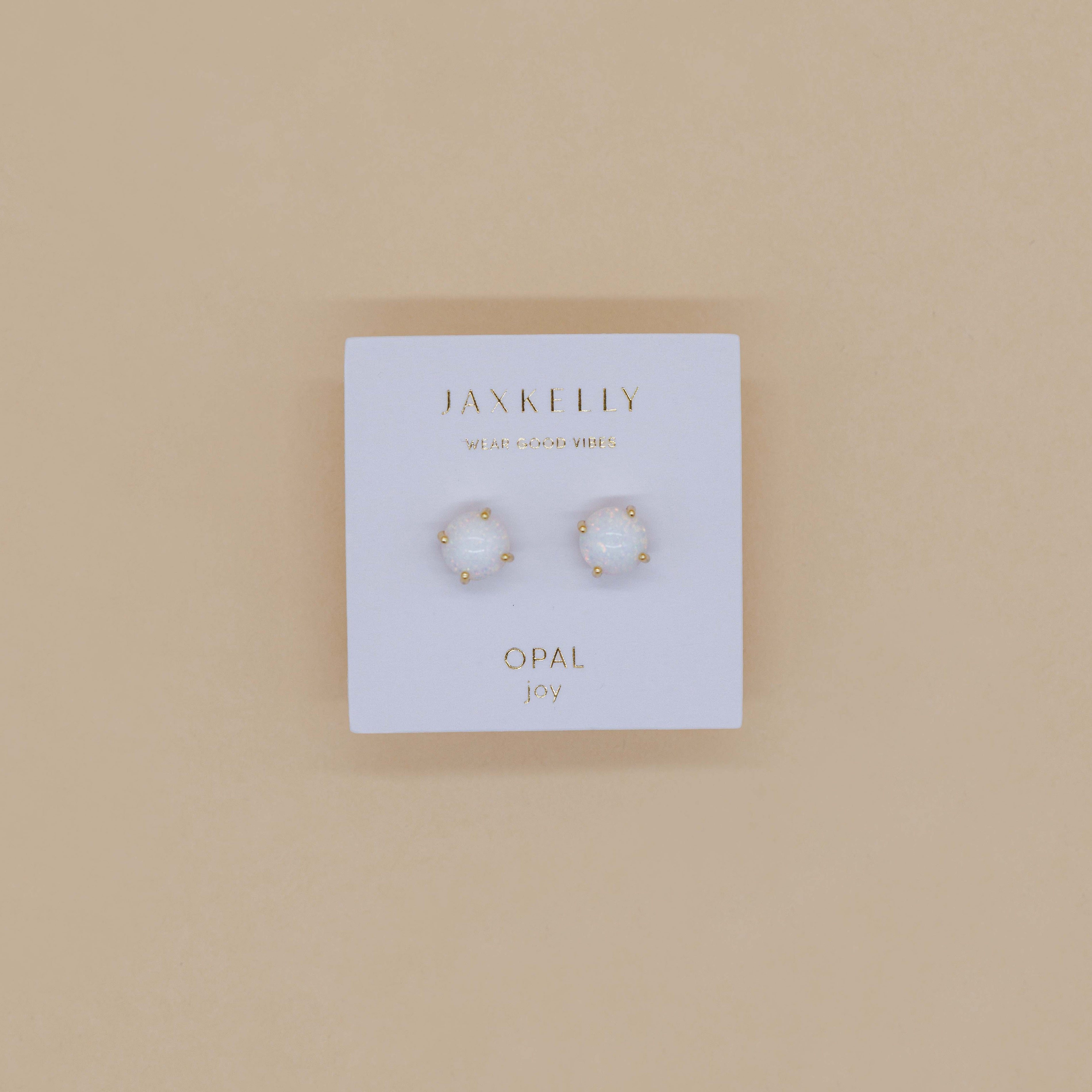 A pair of Round - White Opal Gemstone - Gold Earrings sits on a white Jax Kelly “Opal joy” card against a beige background.