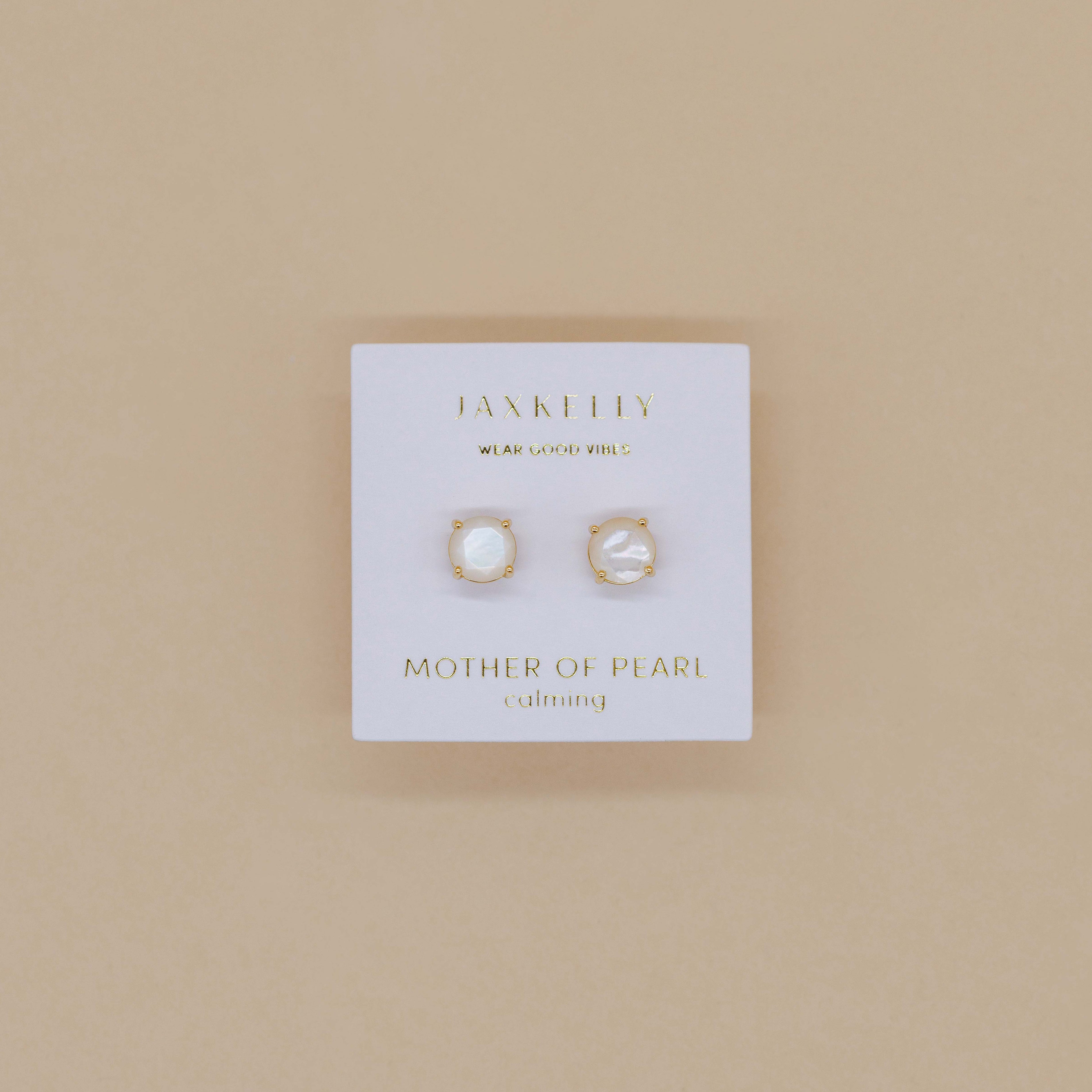 A pair of Round - Mother of Pearl Gemstone - Gold Earrings displayed on a white card labeled JaxKelly – Mother of Pearl – calming, set against a beige background.