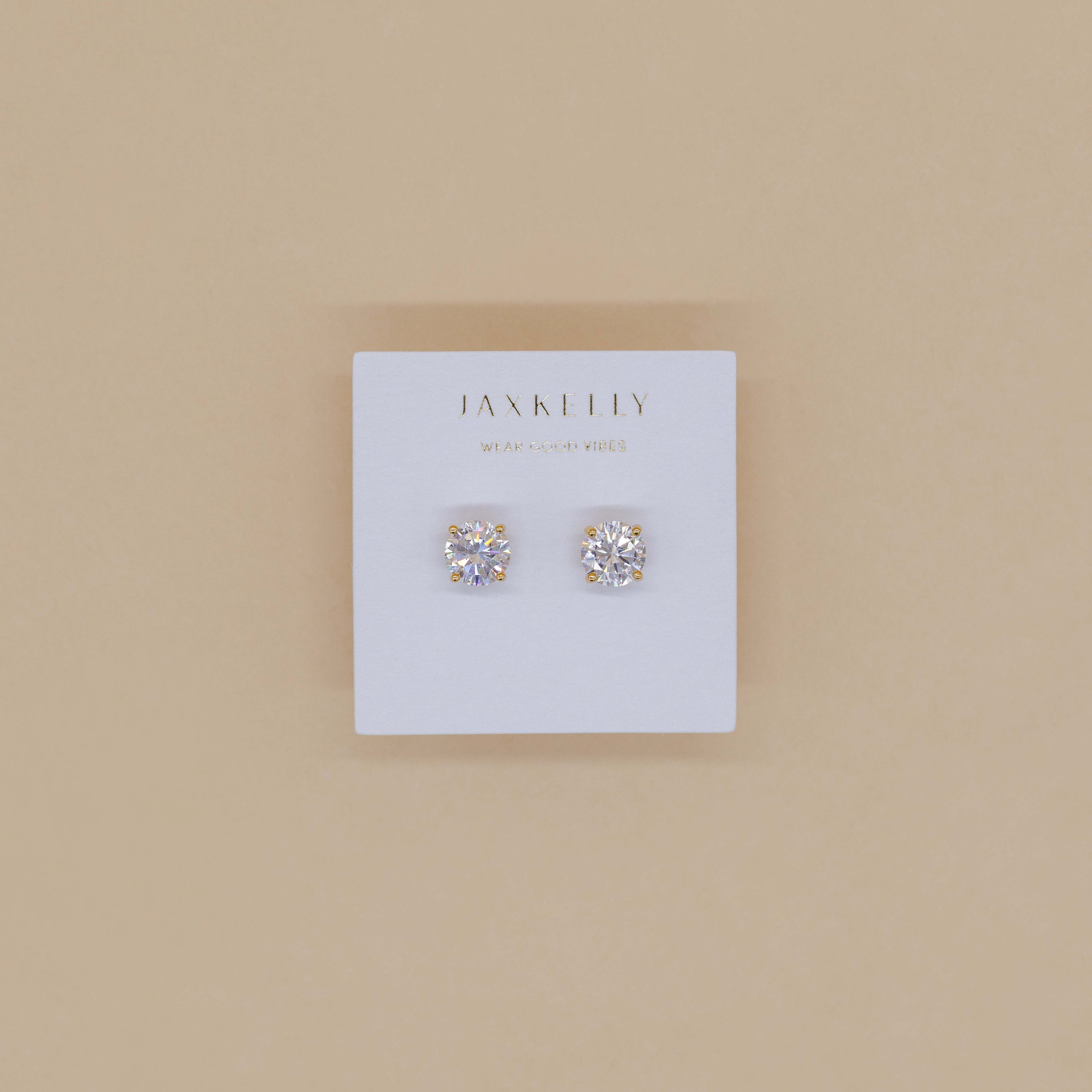 The Round CZ - Clear - Gold Earrings, featuring minimalist gold-plated studs with clear stones, are shown on a white JaxKelly jewelry card against a light beige background.