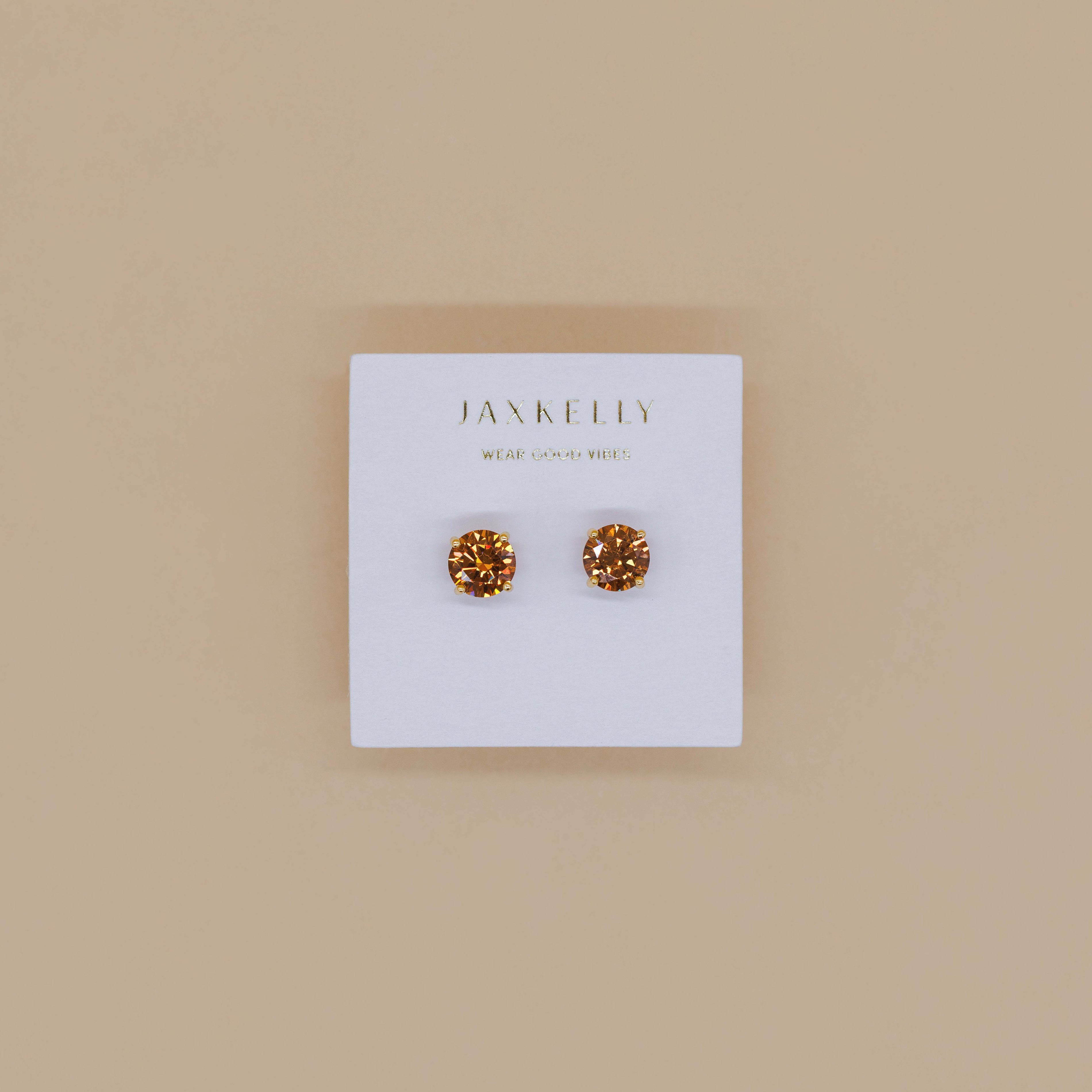 A pair of Round CZ - Coffee - Gold Earrings with a minimalist design is displayed on a white card featuring the JaxKelly brand name, set against a beige background.