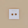A pair of Round CZ - Coffee - Gold Earrings with a minimalist design is displayed on a white card featuring the JaxKelly brand name, set against a beige background.