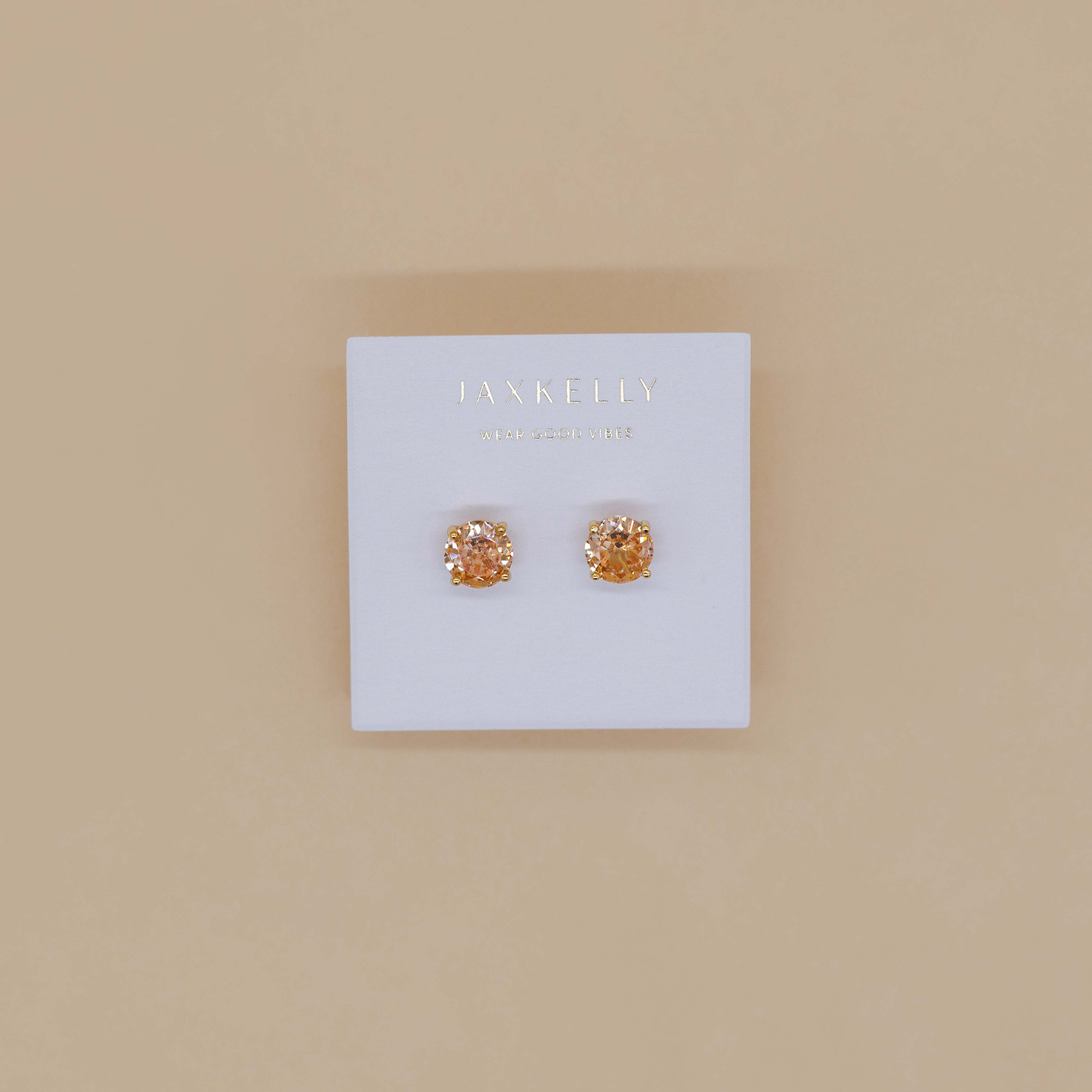 Displayed on a white JaxKelly card against a beige background, the Round CZ- Champagne - Gold Earrings are minimalist gold-plated studs featuring sparkling champagne-colored stones.