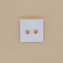 Displayed on a white JaxKelly card against a beige background, the Round CZ- Champagne - Gold Earrings are minimalist gold-plated studs featuring sparkling champagne-colored stones.