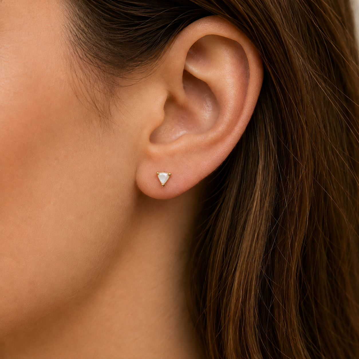 Close-up of a person’s ear adorned with JaxKelly's Mother of Pearl Mini Energy Gems - Calming stud, featuring a small triangular white stone set in 18k gold plating and sterling silver posts. Brown hair is tucked behind the ear against a neutral background.
