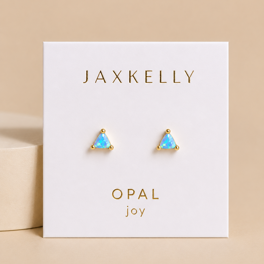 A pair of JaxKelly Fire Opal Mini Energy Gems - Joy studs with gold trim and lab-grown fire opal sit on a white card with gold "JAXKELLY OPAL joy" text, displayed against a beige background.