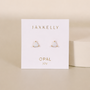 A pair of small, triangular lab-grown White Opal Mini Energy Gems - Joy studs by JaxKelly with 18k gold plating is shown on a white card labeled “JaxKelly” and “Opal joy” in gold, set against a neutral background.